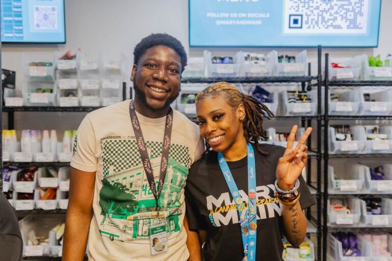 Two smiling people standing close together in an indoor setting with shelves filled with various containers and products in the background. The person on the left is wearing a beige graphic t-shirt and a lanyard with an ID badge, while the person on the right has blond dreadlocks, is wearing a black t-shirt with colorful text, multiple bracelets, and is making a peace sign with their right hand. Both appear happy and friendly.