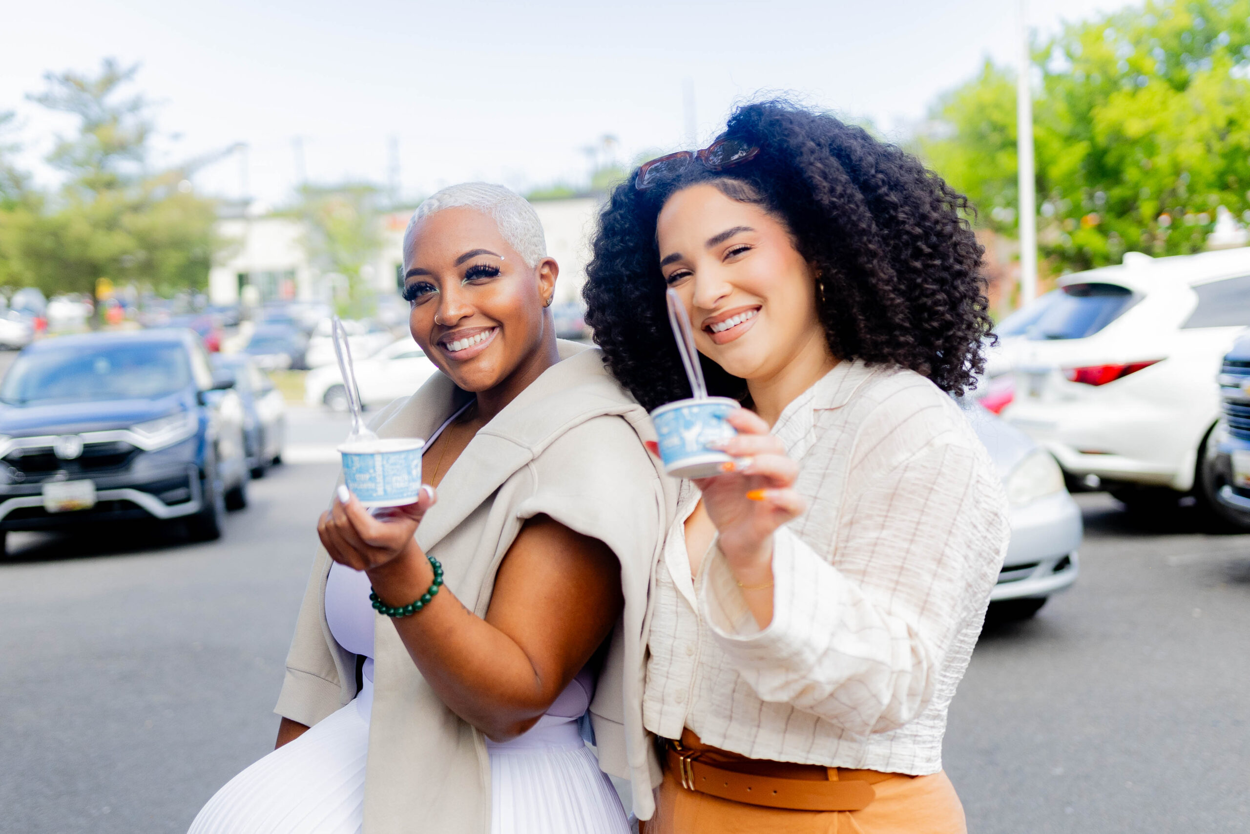 Two smiling women holding cups of ice cream while standing outdoors in a parking lot