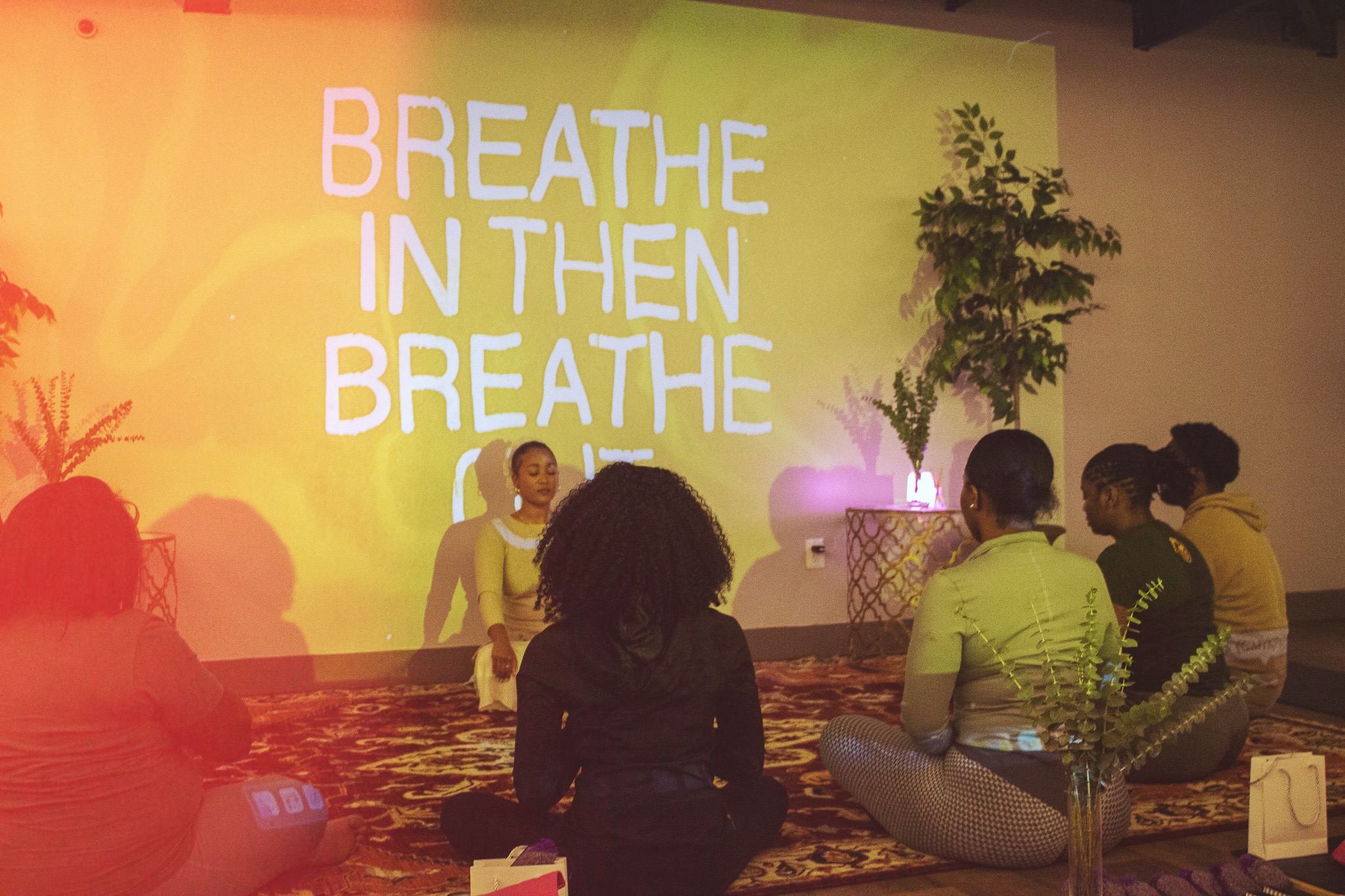 Instructor leading a group meditation session with participants seated on mats in front of a wall projection that reads ‘Breathe In Then Breathe Out