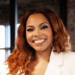 Smiling woman with wavy auburn hair wearing a white blazer in a bright modern office setting
